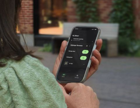 A woman seen from behind holds a smartphone displaying the Parqio app home screen with garage gate and swing door access buttons for Solenga Terrasse.