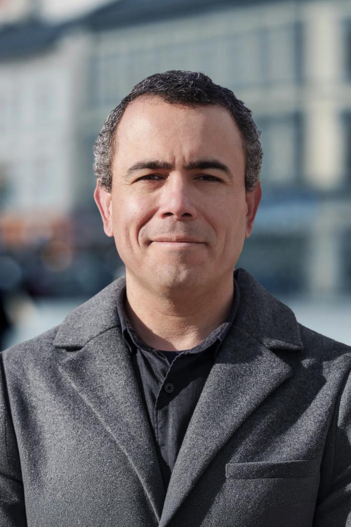 Middle-aged person with short curly hair and a neutral expression stands outdoors wearing a gray coat, with a blurred cityscape in the background.
