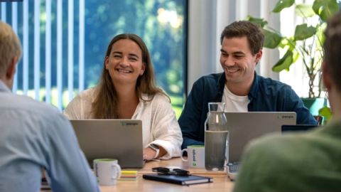 Two smiling colleagues sit at a meeting table with Parqio-branded laptops and mugs, engaged in a team discussion in a bright office.
