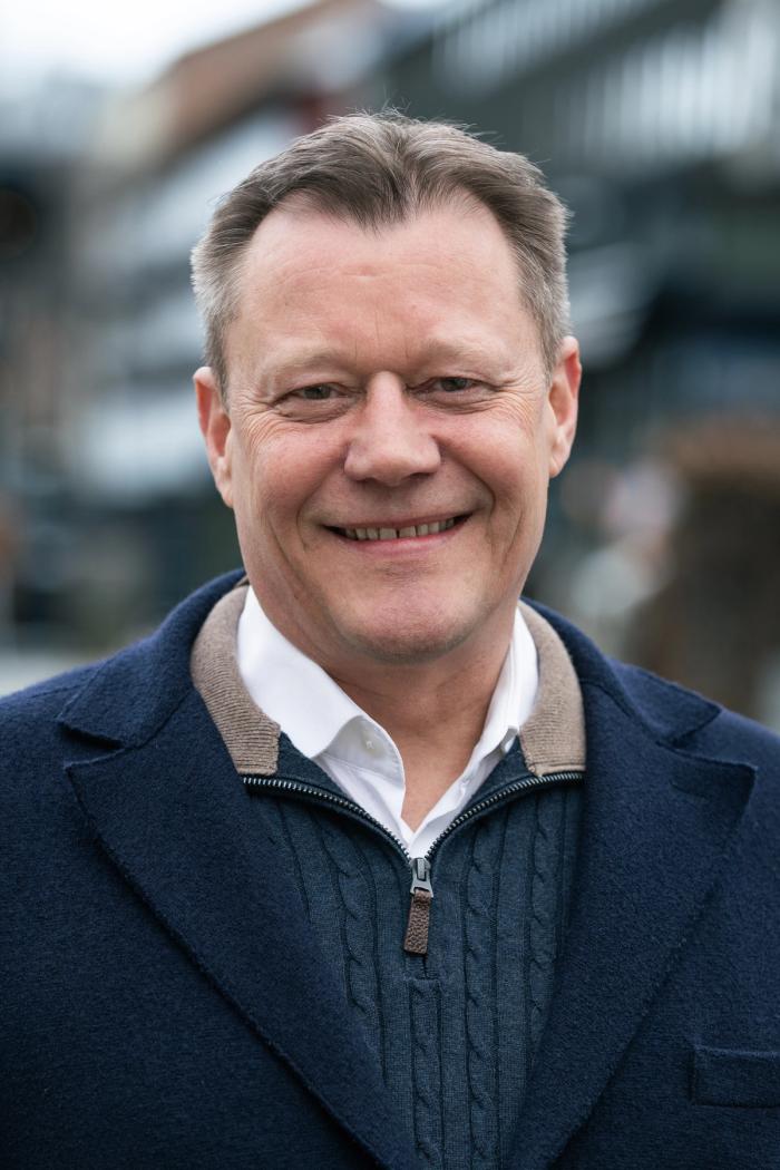 Middle-aged man smiling at the camera outdoors, wearing a navy coat over a zip-up sweater and white collared shirt.