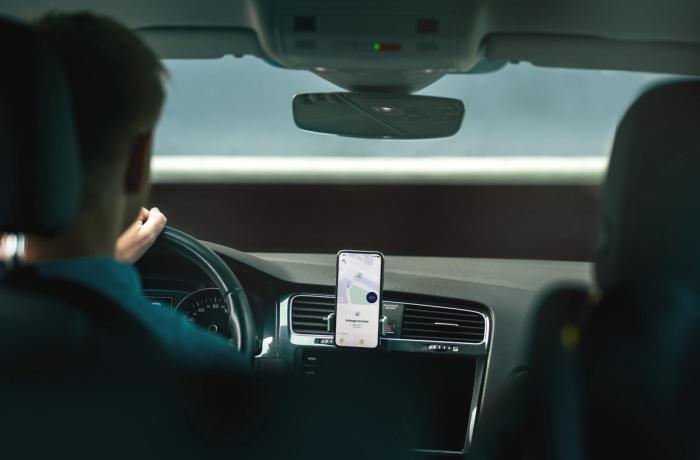 Person driving a car with a smartphone mounted on the dashboard displaying a navigation map.