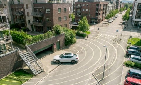 Aerial view of a modern residential complex with a white car driving near a below-ground garage entrance on a curved road lined with parked cars and apartment buildings.
