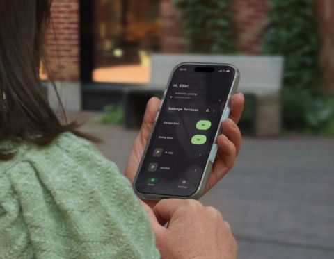 A woman seen from behind holds a smartphone displaying the Parqio app home screen with garage gate and swing door access buttons for Solenga Terrasse.