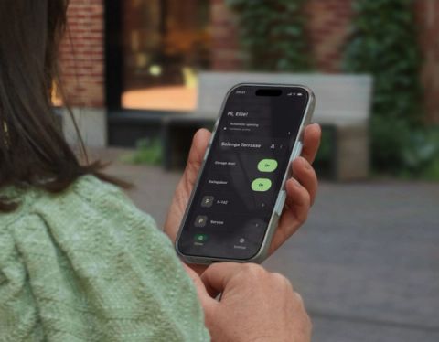 A woman seen from behind holds a smartphone displaying the Parqio app home screen with garage gate and swing door access buttons for Solenga Terrasse.