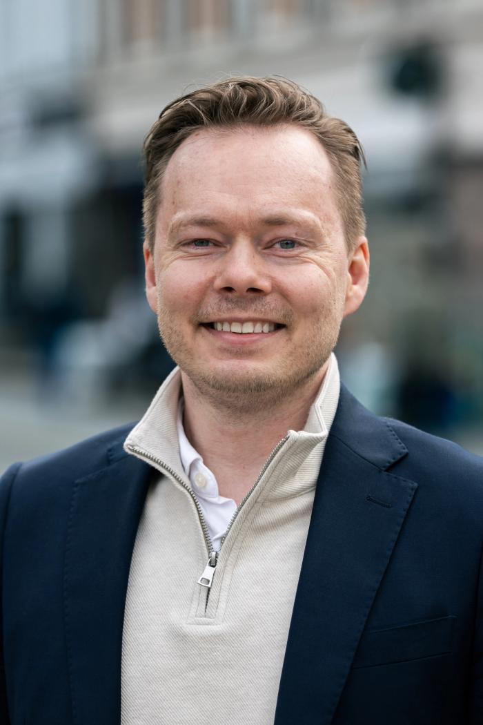 Smiling man in a navy blazer and light zip-up sweater stands outdoors with a blurred city street in the background.