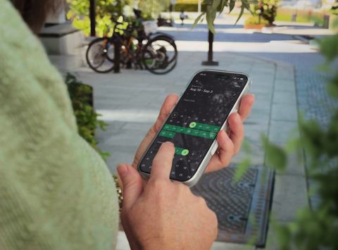 Person in a light green sweater holding a smartphone displaying the Parqio app's date-range calendar picker, with bicycles and a sunny urban footpath visible in the background.