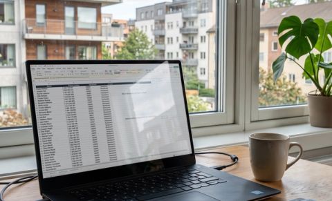 A laptop on a wooden desk displaying a Norwegian-language Excel spreadsheet with resident names, phone numbers, apartment numbers, and access types, with a coffee mug and residential buildings visible through a sunny window in the background.