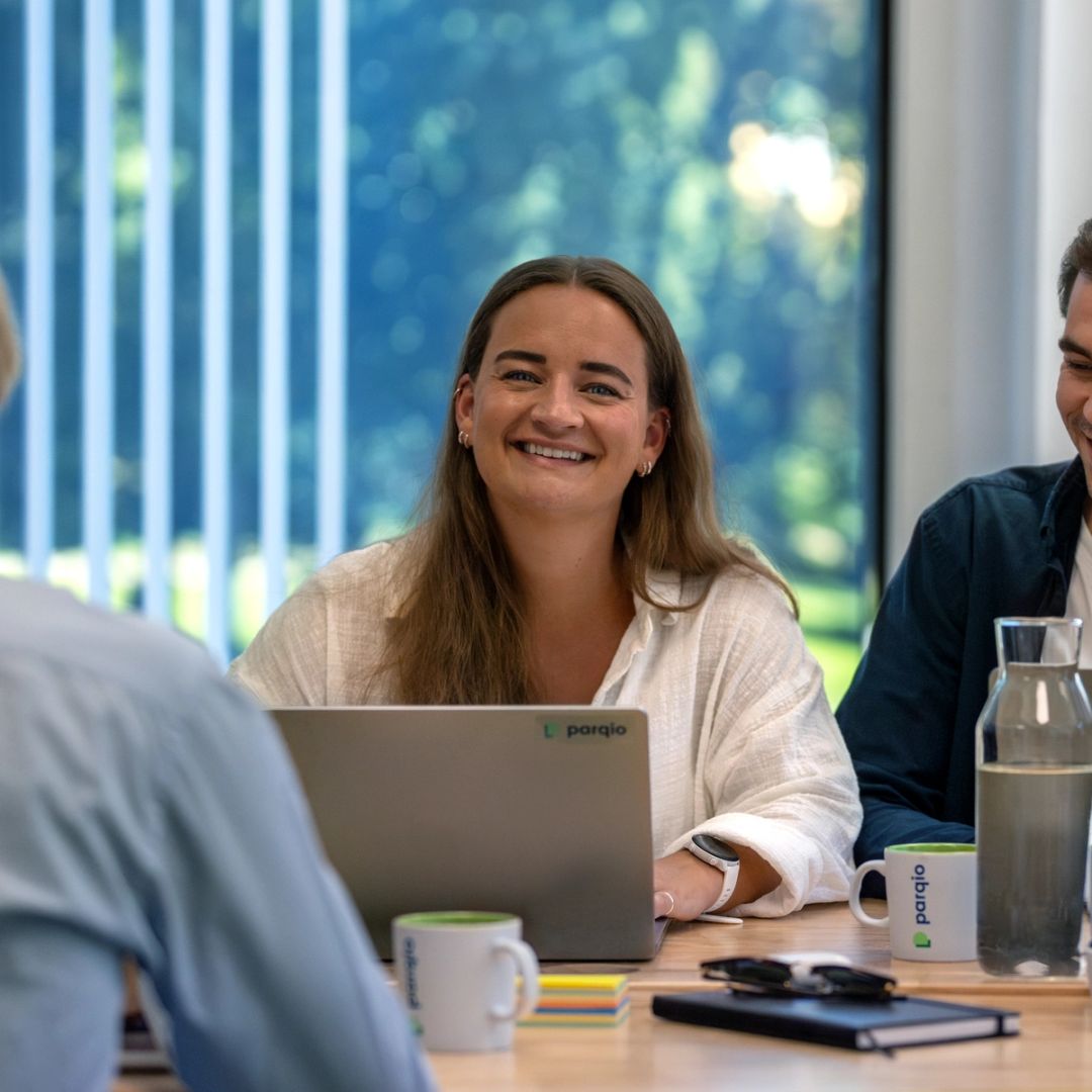 Three smiling colleagues sit around a meeting table with Parqio-branded laptops and mugs during an indoor office meeting.