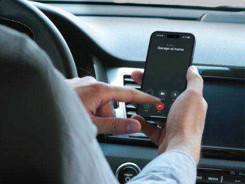 A person sitting in a car holds a smartphone showing an outgoing call screen in Norwegian labelled 'Garasjen hjemme' (Home garage), with a finger about to tap the screen.