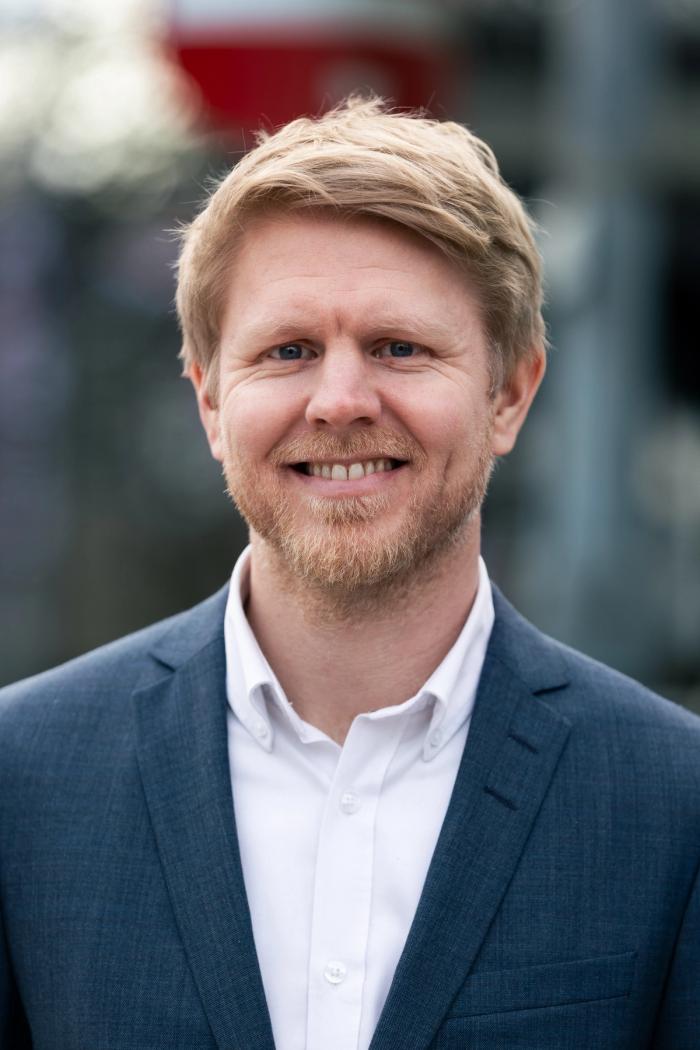 Smiling man with light hair and a short beard wearing a blue suit jacket and white shirt, standing outdoors with a blurred background.
