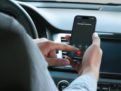 En person sitter i en bil og holder en smarttelefon med en utgående samtale-skjerm på norsk merket 'Garasjen hjemme', med en finger som er i ferd med å trykke på skjermen.