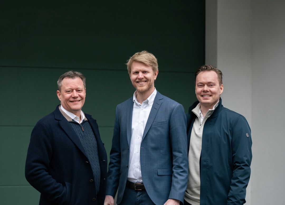 Three men in smart casual business attire stand side by side indoors, facing the camera and smiling slightly.