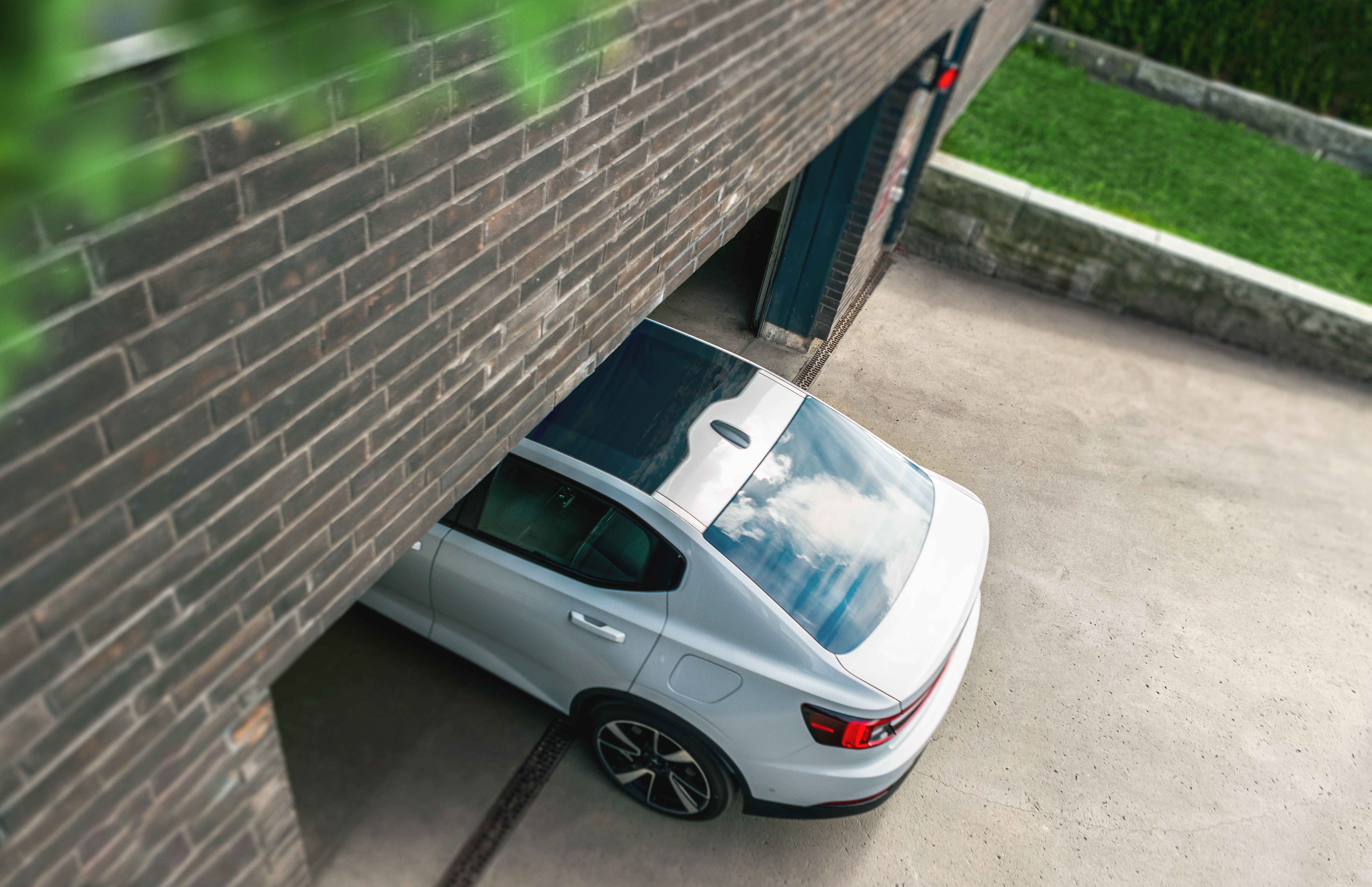 Aerial view of a white electric sedan entering a residential parking area through a gate beside a brick building, with a concrete driveway and green lawn visible.