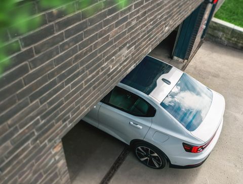 Aerial view of a white electric sedan entering a residential parking area through a gate beside a brick building, with a concrete driveway and green lawn visible.