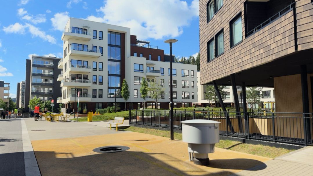 Modern apartment buildings with balconies line a sunny pedestrian street with benches, greenery, and a few people walking and cycling.