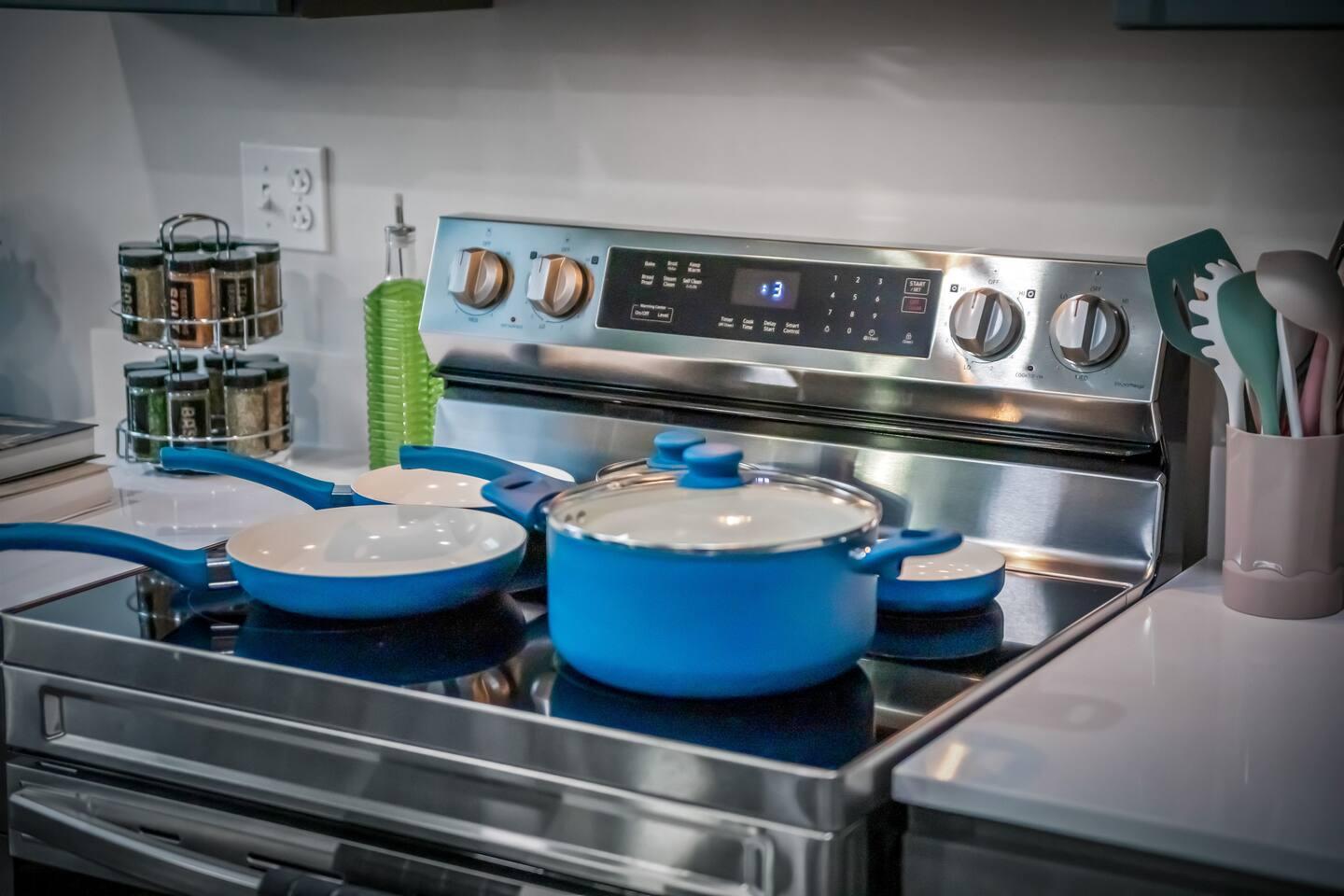 Kitchen area with electric stove, pots, pans, spices, oil, ladles and an electric socket.