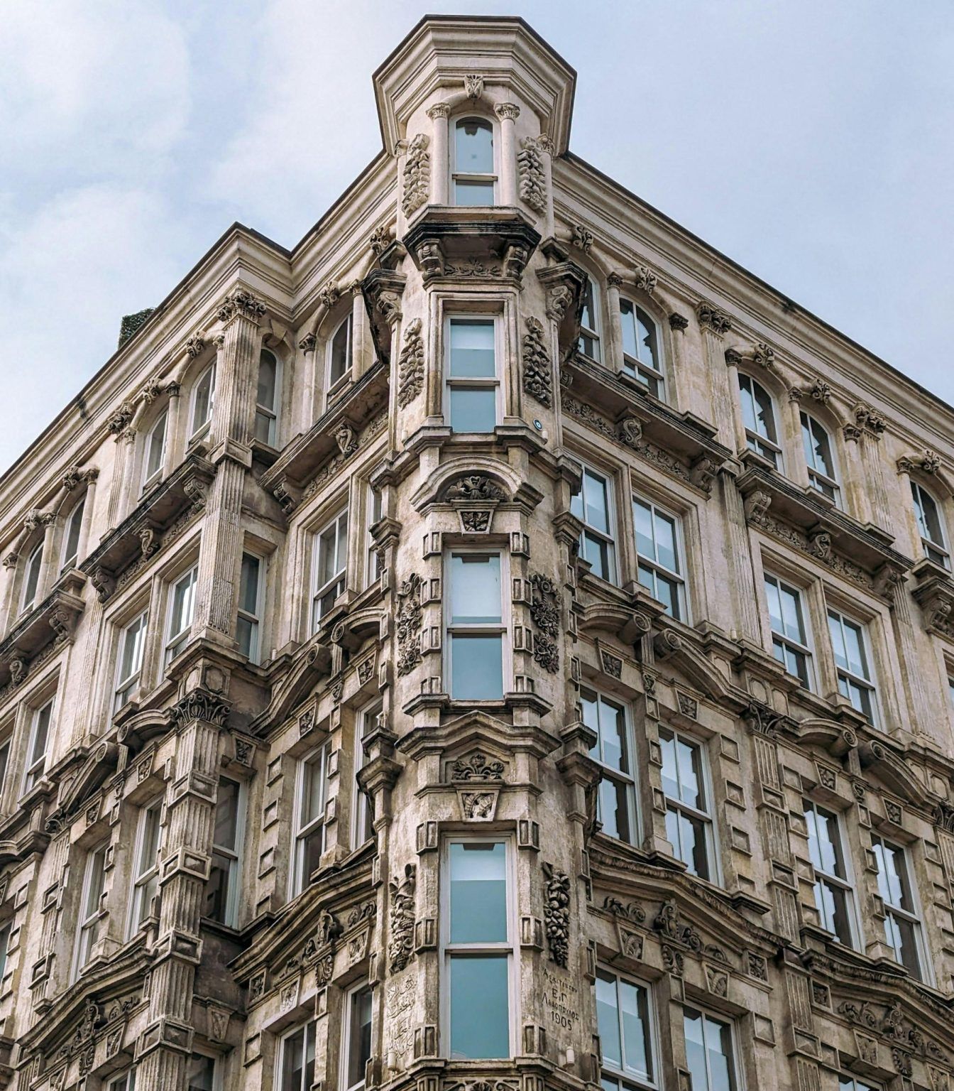 Ein historisches, reich verziertes Gebäude mit detaillierten Steinmetzarbeiten und hohen, schmalen Fenstern auf jeder Etage, aus der Eckperspektive vor einem bewölkten Himmel betrachtet.