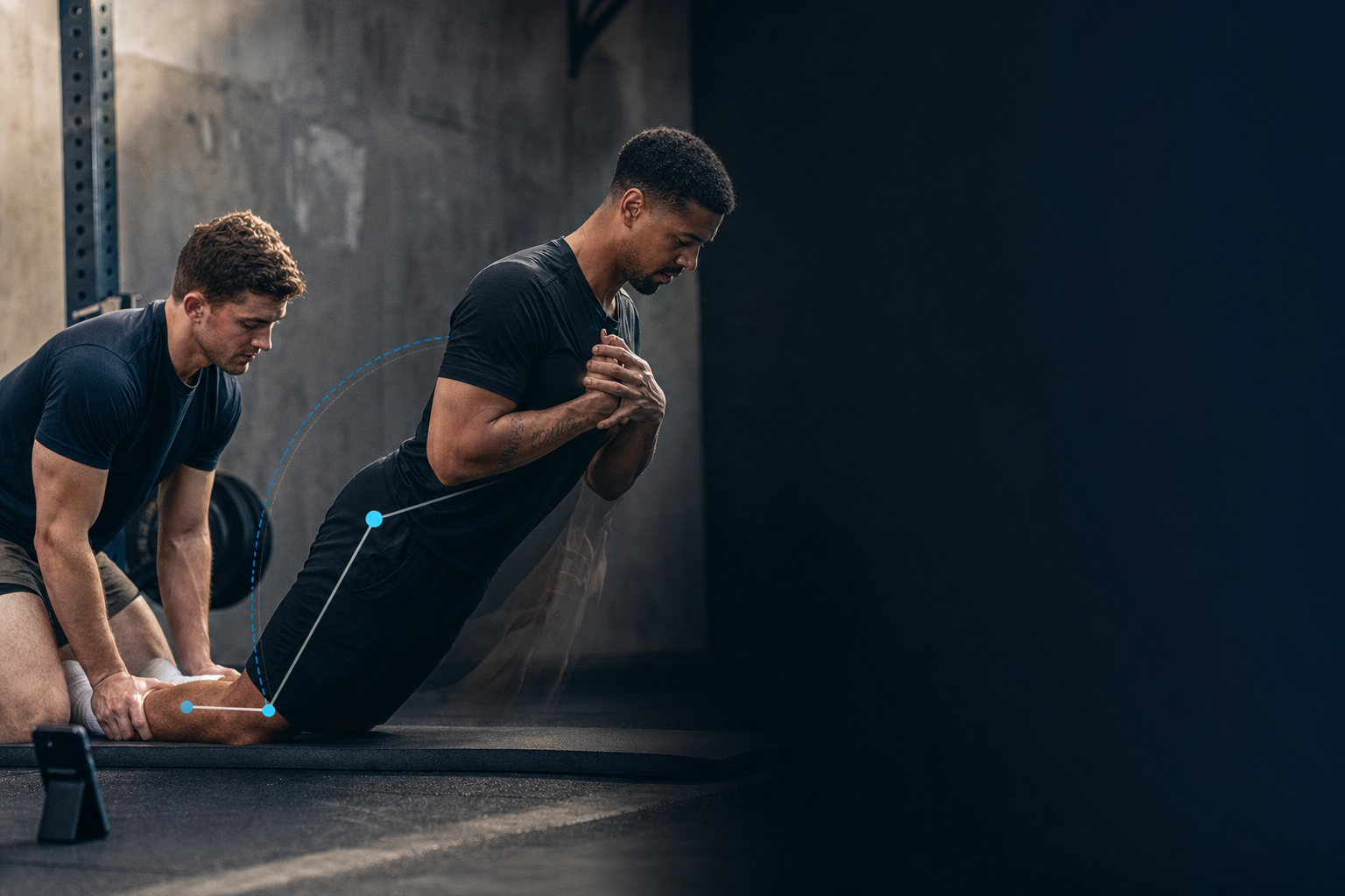 Athlete lowering into a Nordic curl on a mat while a training partner anchors the ankles and a phone films the descent from the side