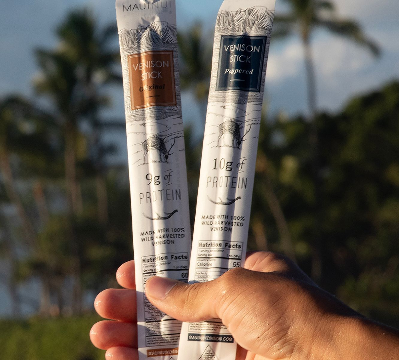A hand holding up two Maui Nui Venison sticks against a backdrop of palm trees and blue sky — one "Original" flavor with 9g of protein, one "Peppered" flavor with 10g of protein, each labeled "Made with 100% Wild Harvested Venison."