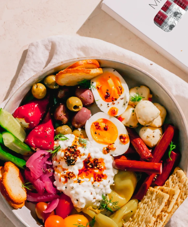 A colorful snack bowl featuring Maui Nui Venison meat sticks alongside soft-boiled eggs, cottage cheese, olives, pickled vegetables, cherry tomatoes, cucumbers, and crackers, garnished with fresh dill and chili flakes.
