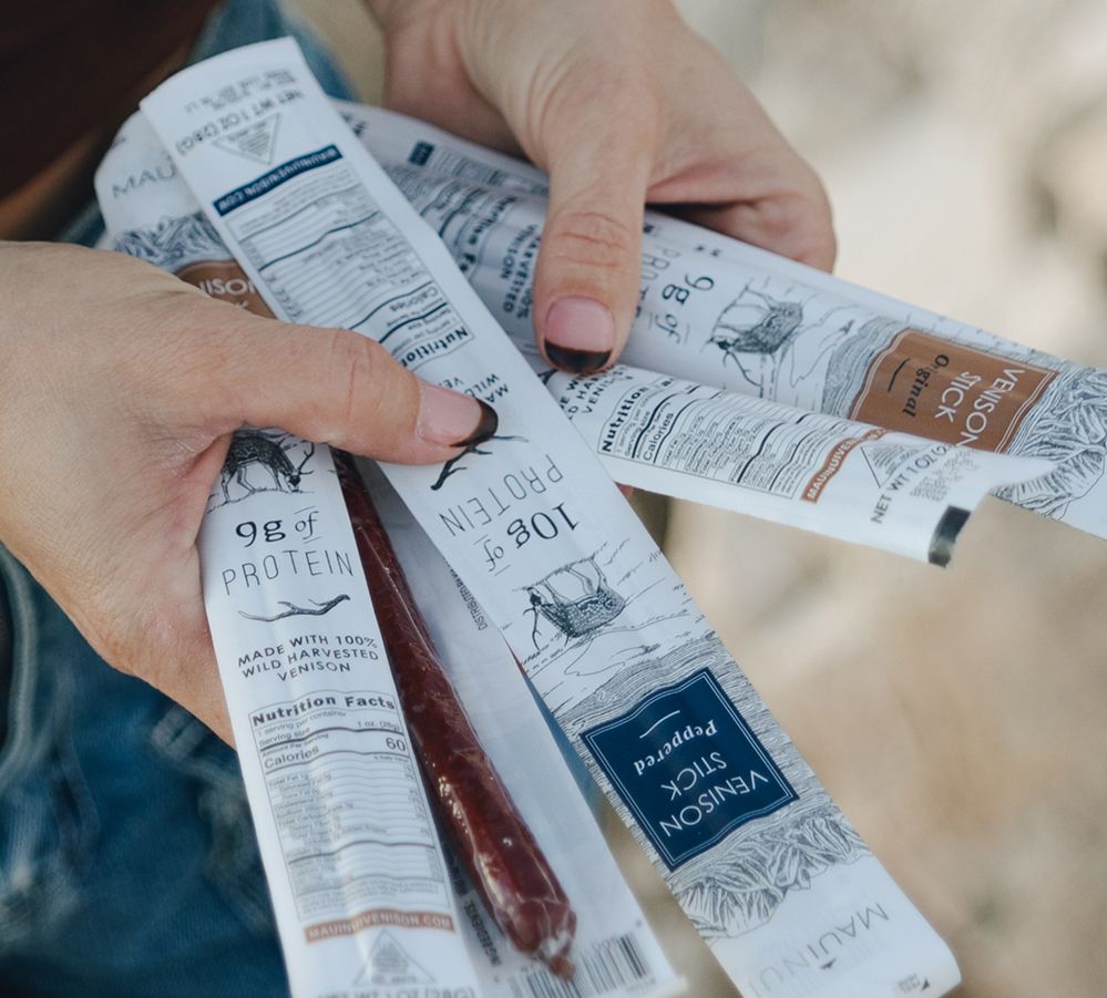 Close-up of two hands holding several Maui Nui Venison stick packages fanned out, with one opened wrapper revealing a dark red venison stick. Packaging text reads "9g of Protein," "Made with 100% Wild Harvested Venison," "Venison Stick Original," and "Venison Stick Peppered," with nutrition facts panels visible.