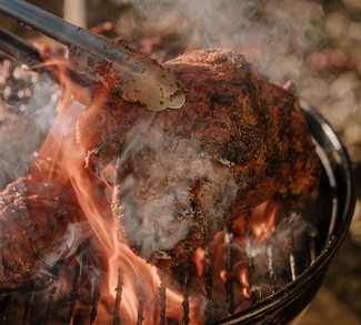 Close-up of a well-seared cut of meat being lifted with tongs off an open-flame grill, smoke rising and flames visible through the charcoal grate.