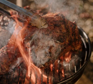 Close-up of a well-seared cut of meat being lifted with tongs off an open-flame grill, smoke rising and flames visible through the charcoal grate.