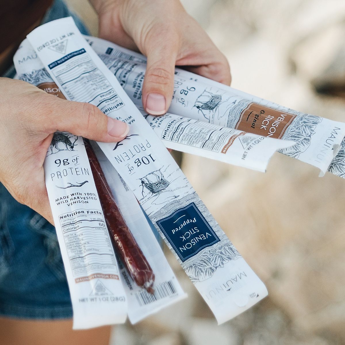 Close-up of two hands holding several Maui Nui Venison stick packages fanned out, with one opened wrapper revealing a dark red venison stick. Packaging text reads "9g of Protein," "Made with 100% Wild Harvested Venison," "Venison Stick Original," and "Venison Stick Peppered," with nutrition facts panels visible.