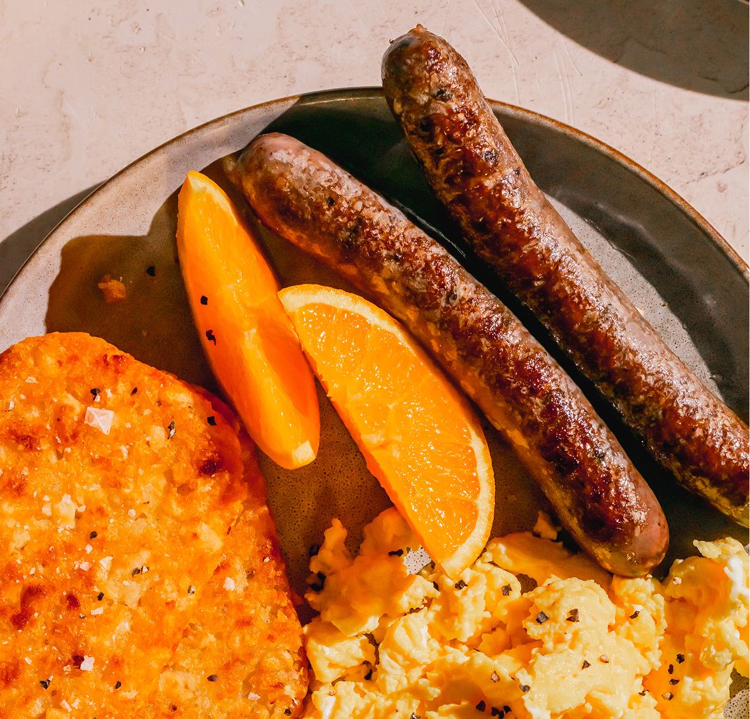 Overhead shot of a breakfast plate with scrambled eggs, a crispy hashbrown, two orange slices, and two grilled Maui Nui breakfast links, set on a white plate.