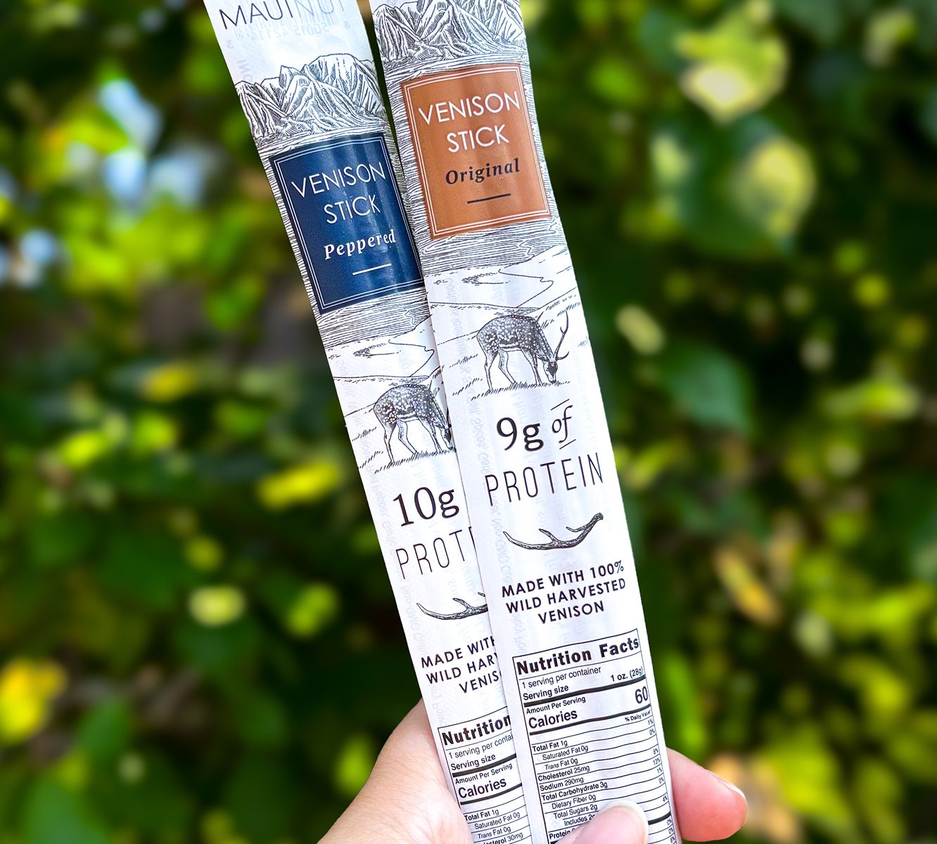 A hand holding up two Maui Nui Venison sticks against a backdrop of green shrubbery — one "Original" flavor with 9g of protein, one "Peppered" flavor with 10g of protein, each labeled "Made with 100% Wild Harvested Venison."