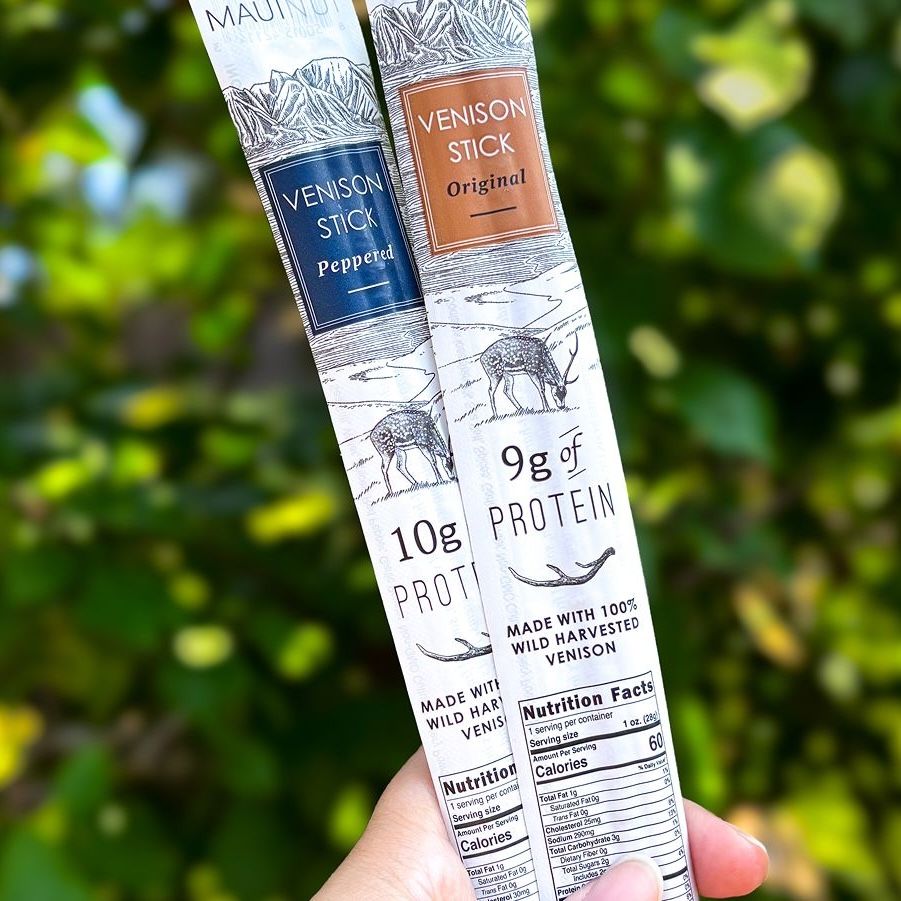 Two Maui Nui Venison Stick packages, Peppered and Original, held fanned out against a blurred green leafy background, showing "10g Protein" and "9g of Protein" labels with visible nutrition facts panels.