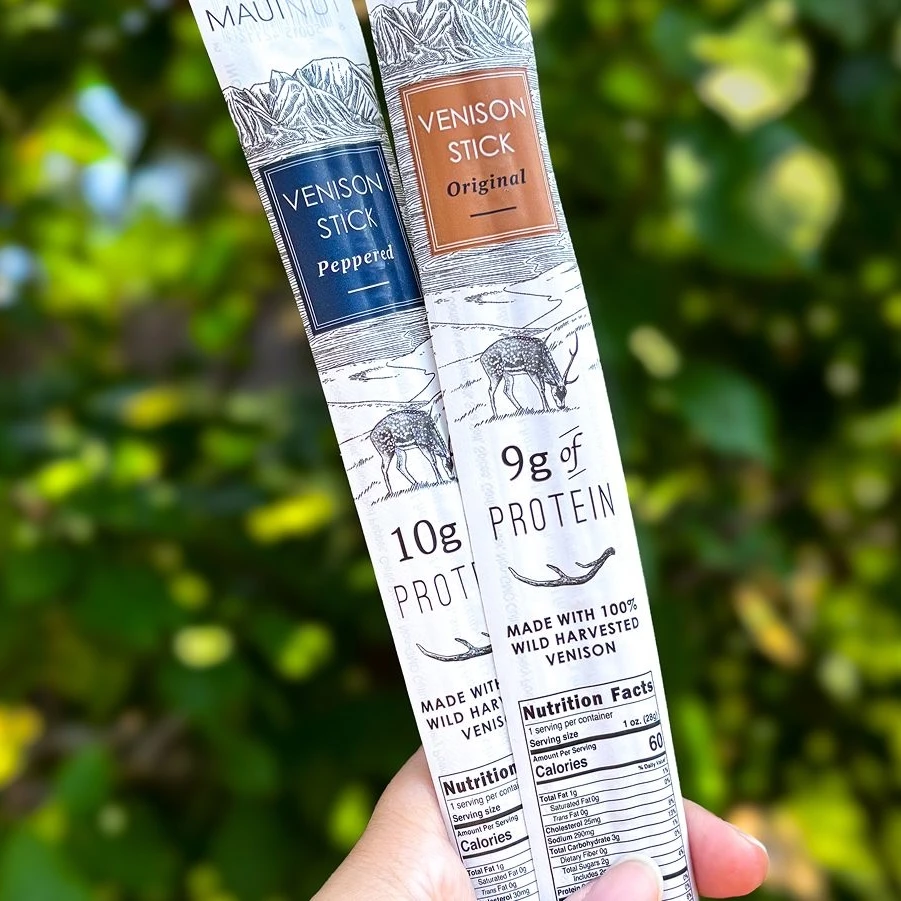 Two Maui Nui Venison Stick packages, Peppered and Original, held fanned out against a blurred green leafy background, showing "10g Protein" and "9g of Protein" labels with visible nutrition facts panels.