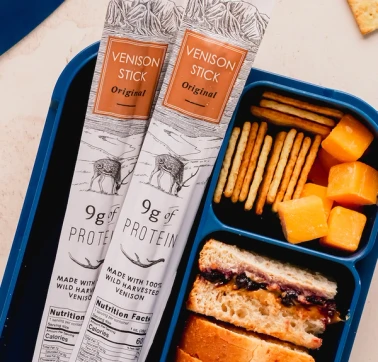 Two Maui Nui Venison Original sticks resting against a blue bento-style lunch box filled with crackers, cheddar cheese cubes, and a PB&J sandwich.