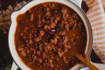Maui Nui ground venison chili in a large pot filled with red kidney beans, ground venison and diced tomatoes with a resting wooden spoon.