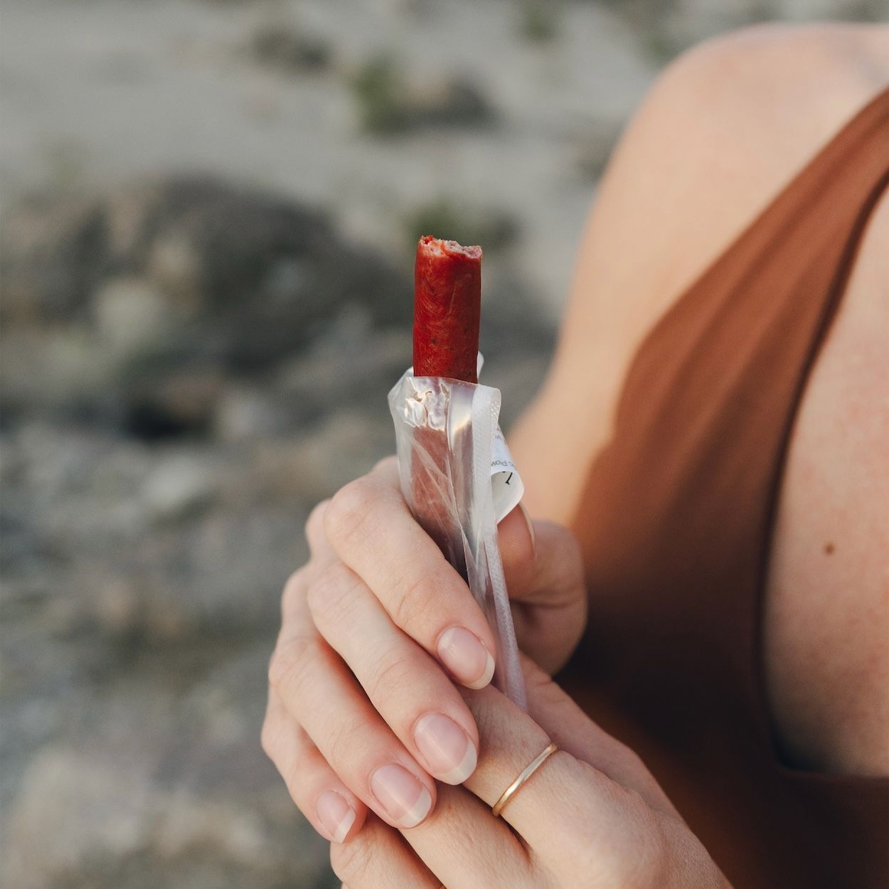 Close-up of a partially eaten Maui Nui Venison stick held upright in its opened wrapper, gripped by a hand with a small arrow tattoo and ombre manicure, outdoors in soft evening light.