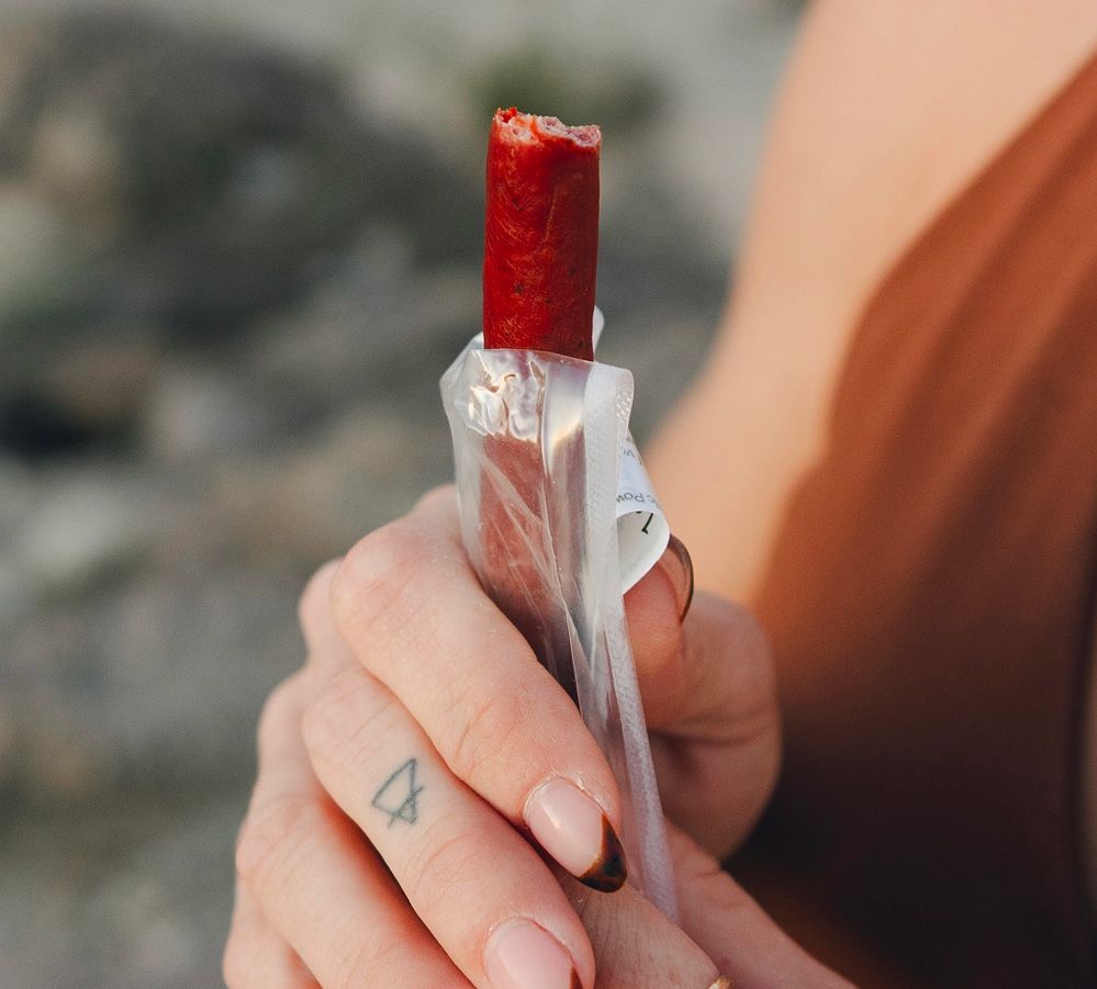 Close-up of a partially eaten Maui Nui Venison stick held upright in its opened wrapper, gripped by a hand with a small arrow tattoo and ombre manicure, outdoors in soft evening light.
