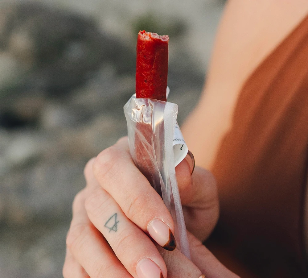 Close-up of a partially eaten Maui Nui Venison stick held upright in its opened wrapper, gripped by a hand with a small arrow tattoo and ombre manicure, outdoors in soft evening light.