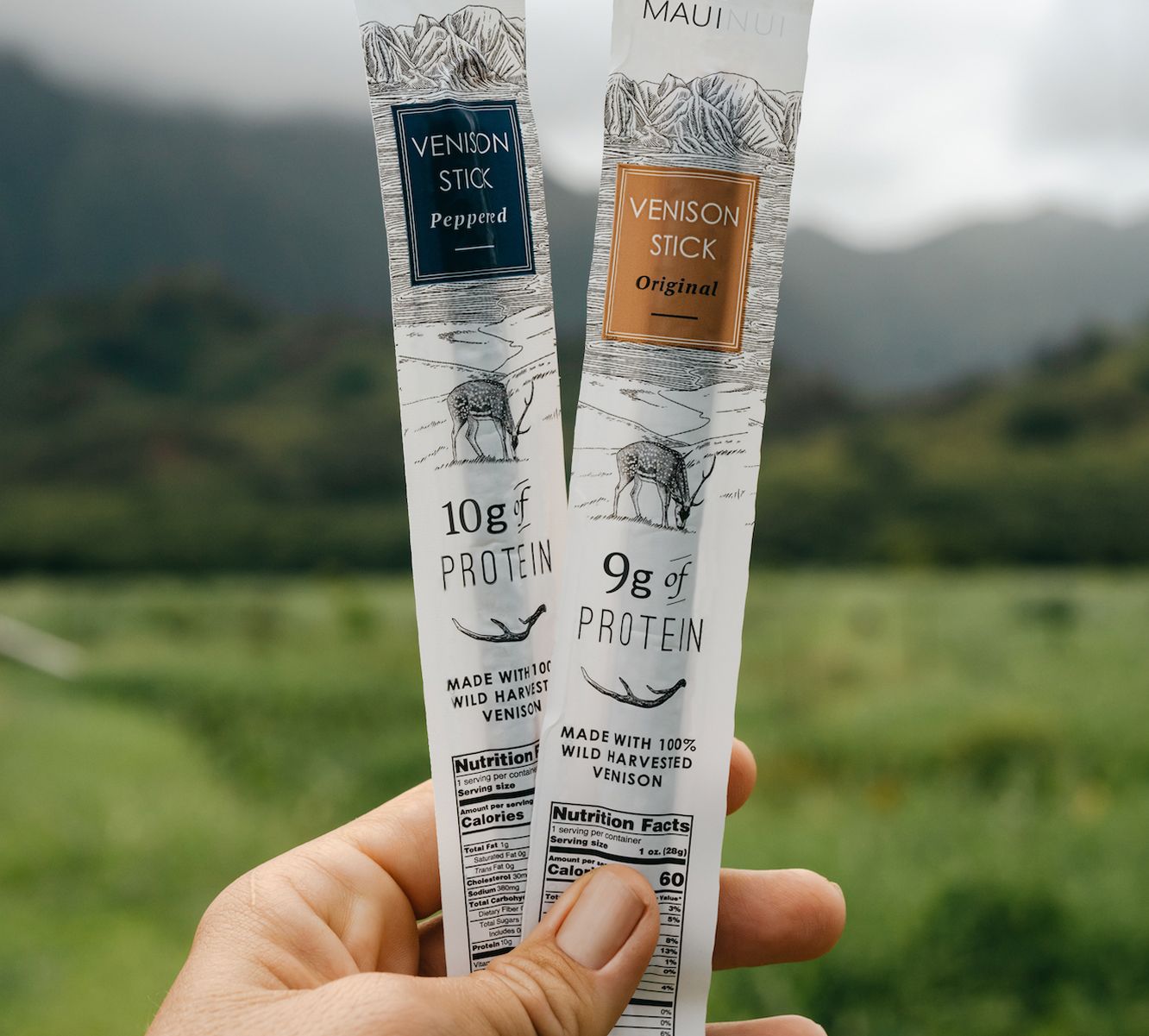 A hand holding up two Maui Nui Venison sticks against a backdrop of green pastures and mountains — one "Original" flavor with 9g of protein, one "Peppered" flavor with 10g of protein, each labeled "Made with 100% Wild Harvested Venison."