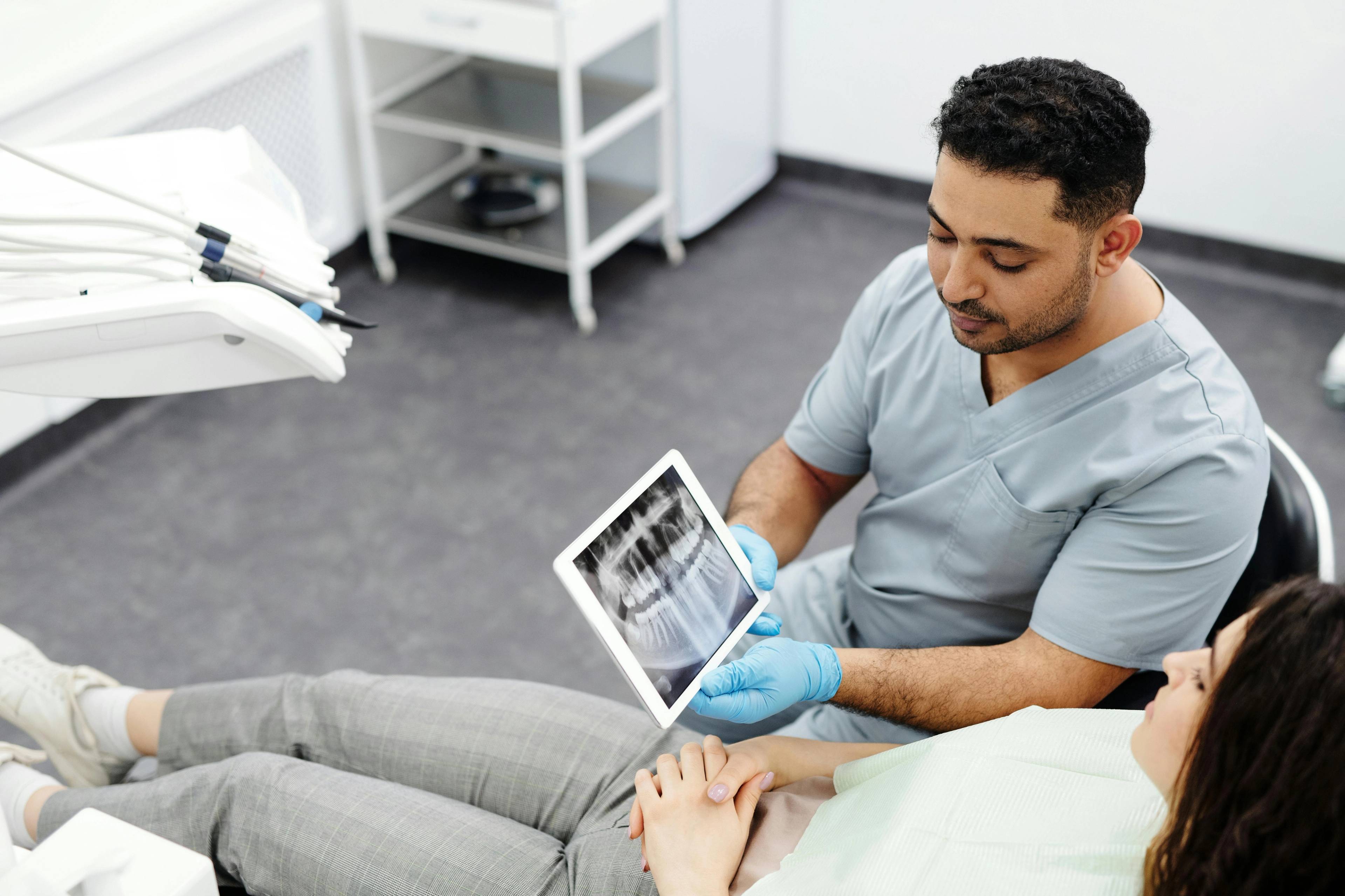 dentist in blue gloves holding screen showing seated patient an x-ray of teeth in surgical environment
