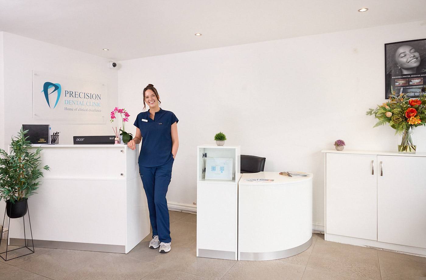 Inside precision dental clinic patient waiting area and reception dental nurse smiling to welcome patients featuring clean floor and chairs a large window some houseplants and brand signage