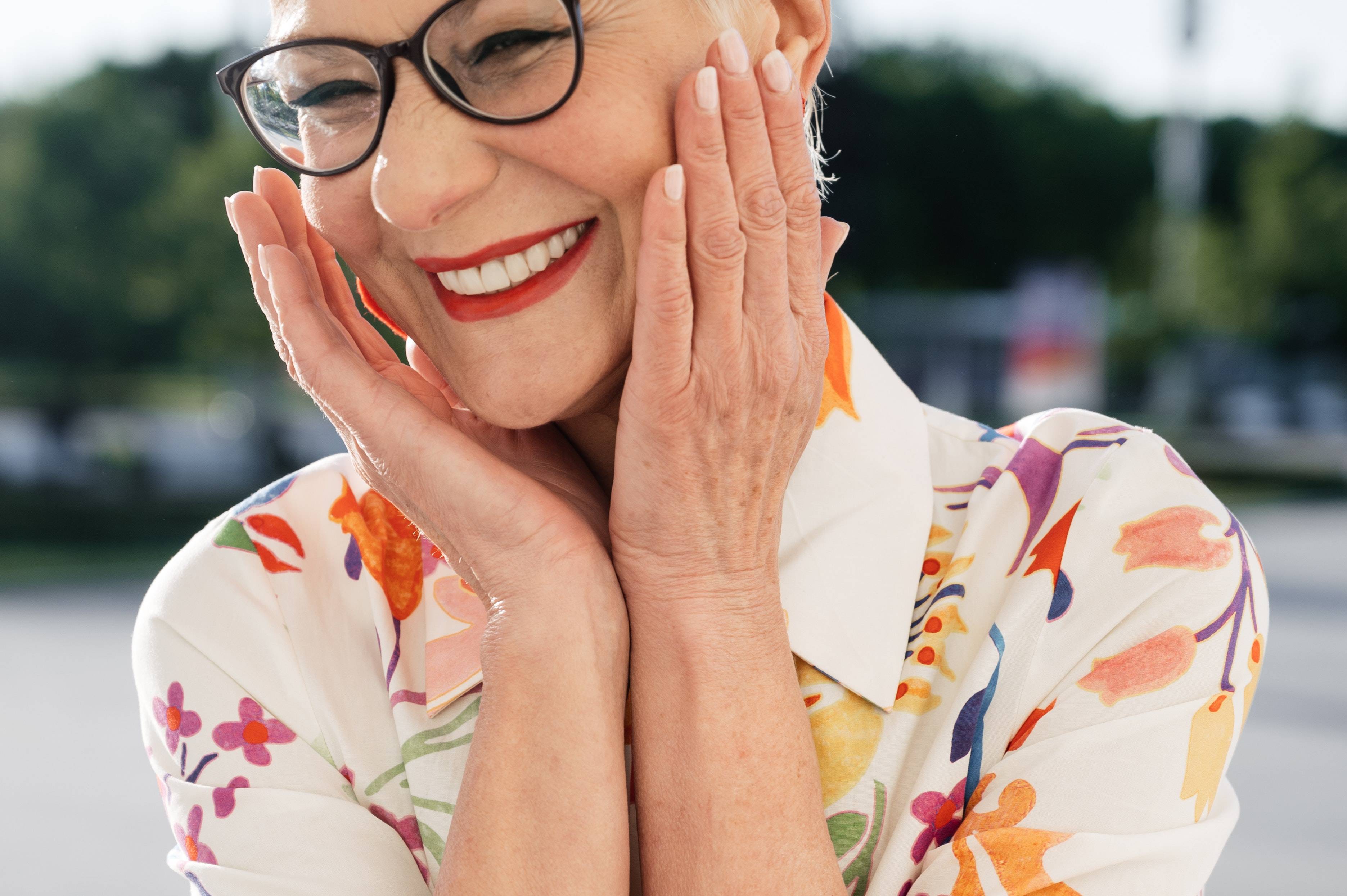 Female patient smiling outside in the sun wearing black framed spectacles bright red lips and a colourful shirt