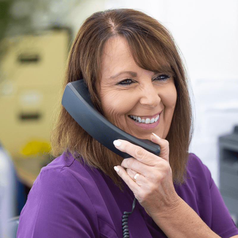 Female receptionist with brown hair and purple uniform smiling and holding a phone up to ear