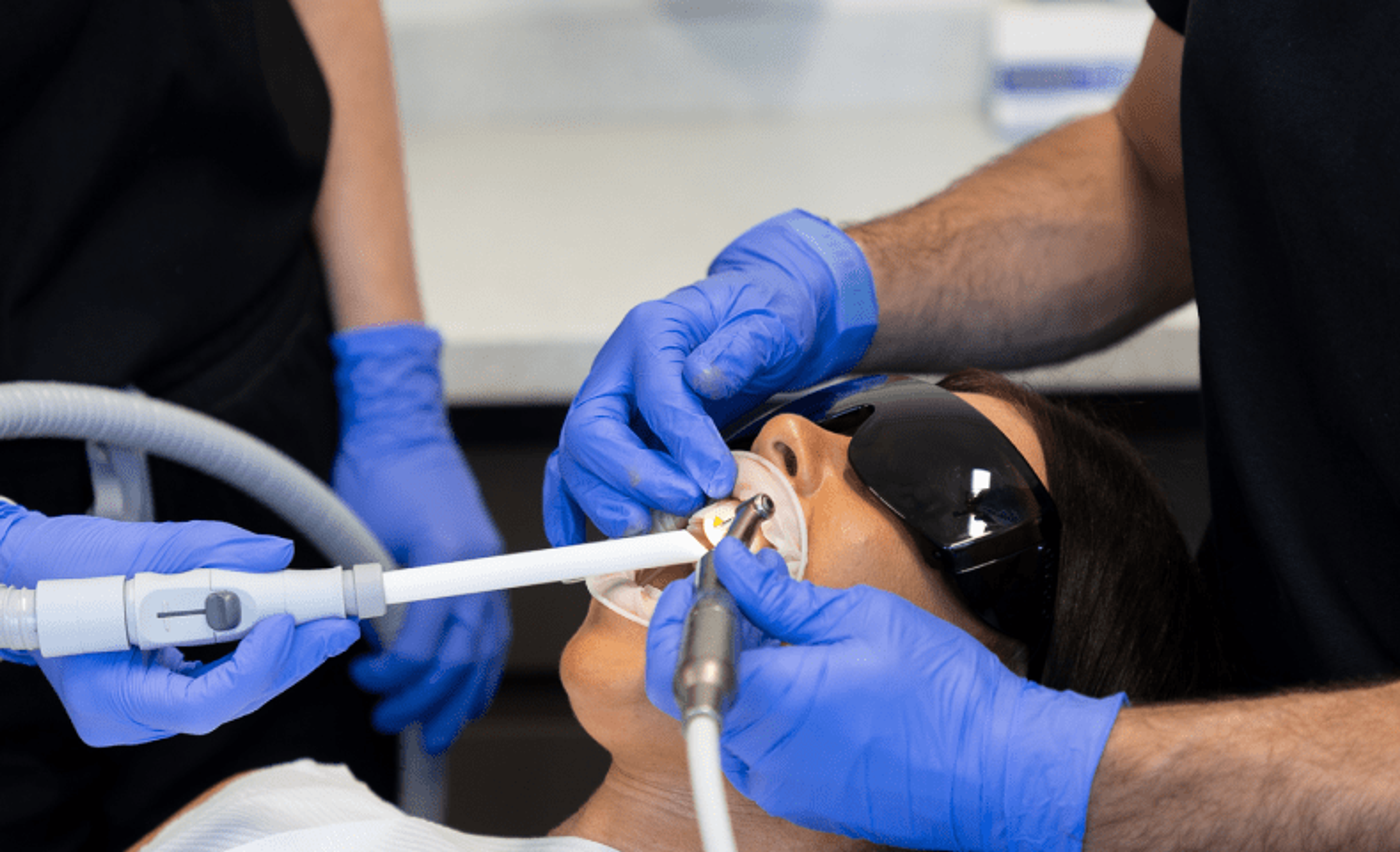 dentist wearing blue gloves holding dental implements inside patients mouth while patient wears eye protection