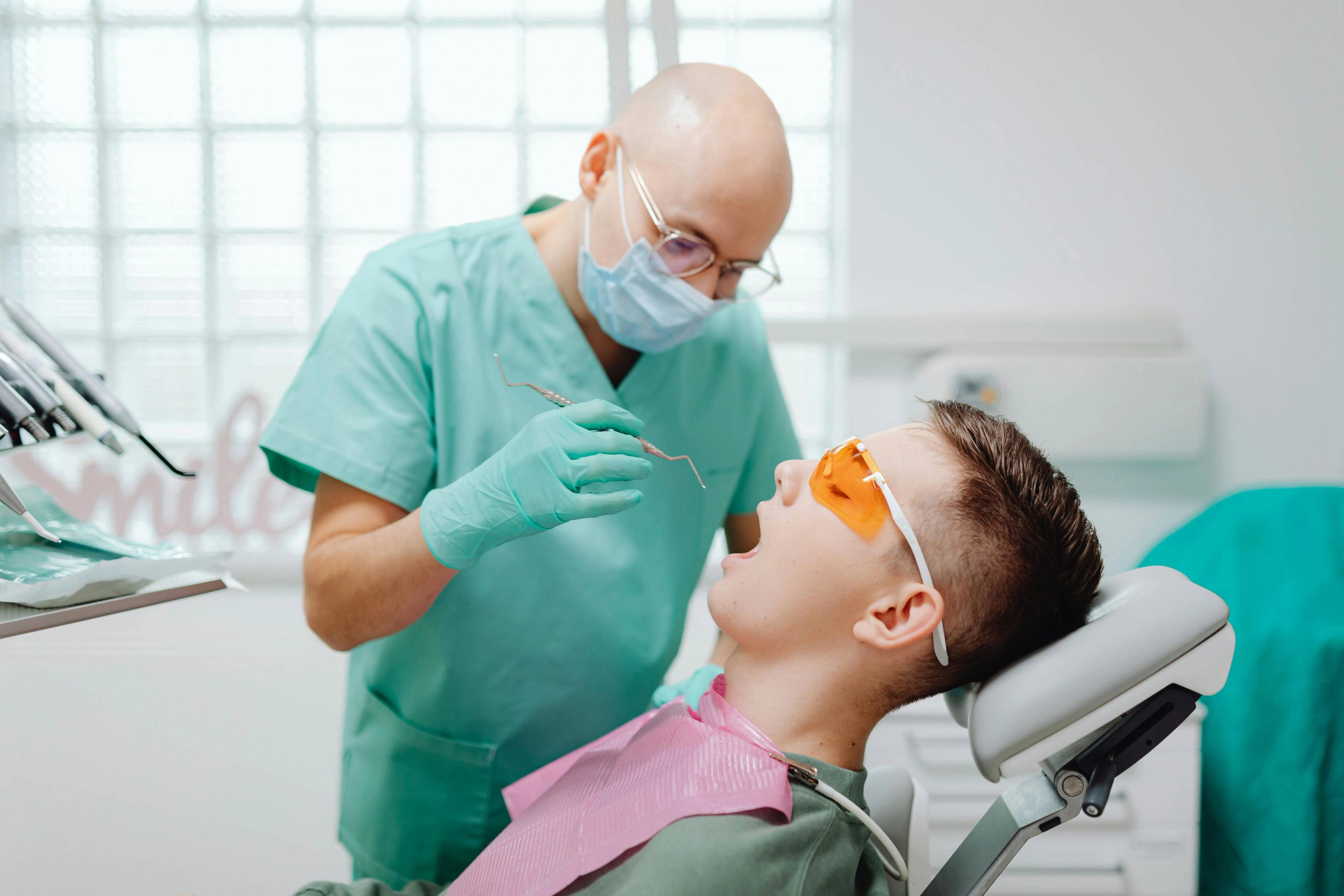 dentist wearing blue uniform gloves and face mask holding dental tools inside young male patient mouth while seated wearing protective glasses