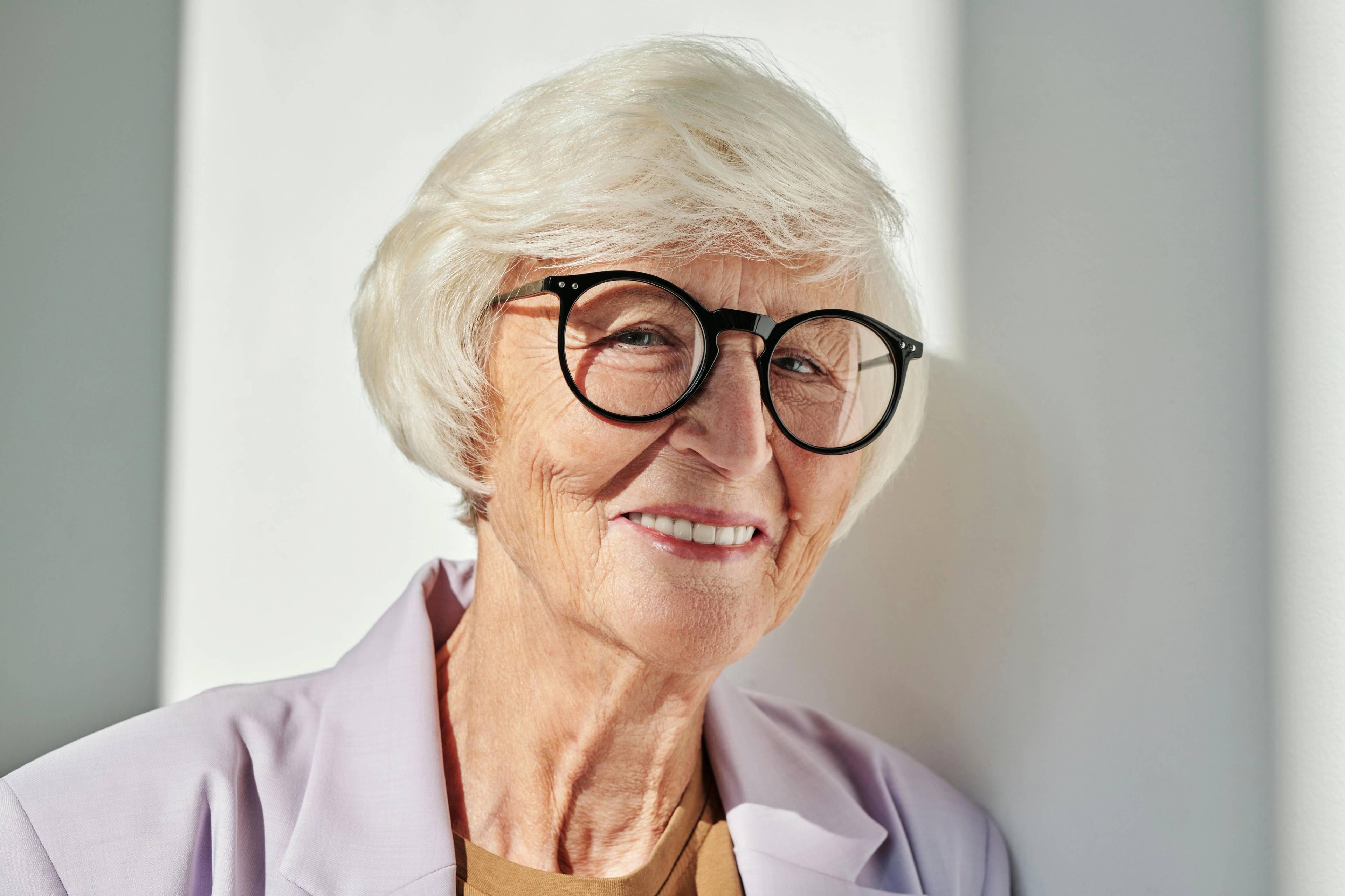 elderly female patient with white hair wearing thick black glasses and a lilac shirt smiling in the sun