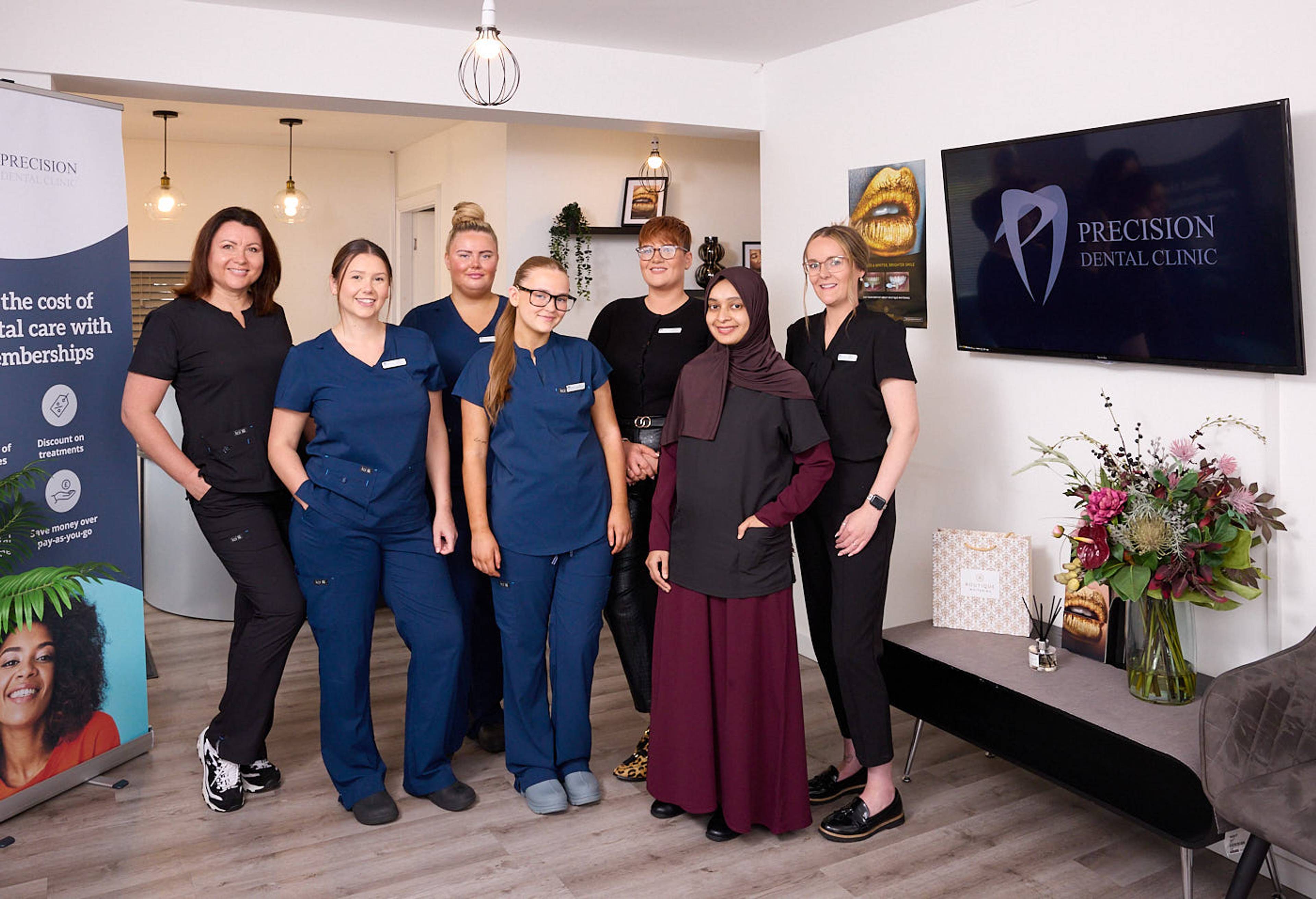 team photo at precision dental care featuring seven dental professionals smiling in patient waiting area under warm lighting with brand signage on screen
