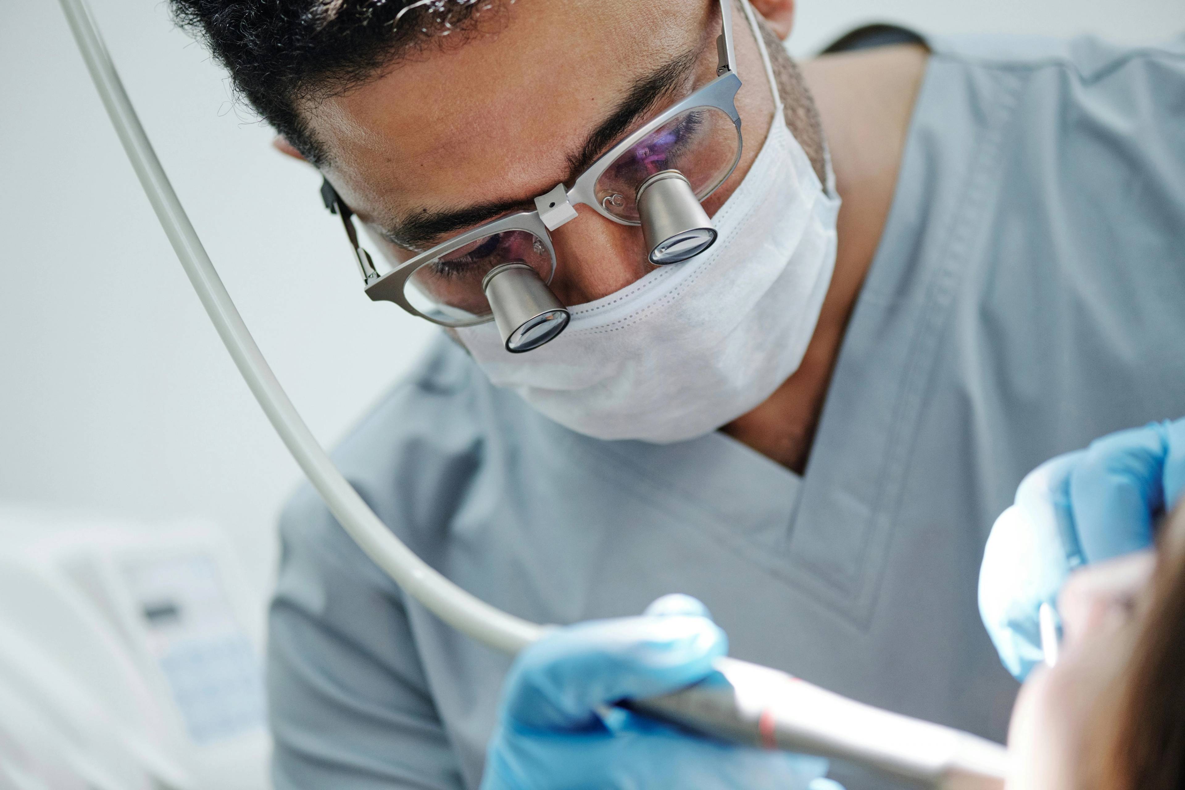 dentist wearing blue uniform gloves and loupes holding dental equipment in patients mouth