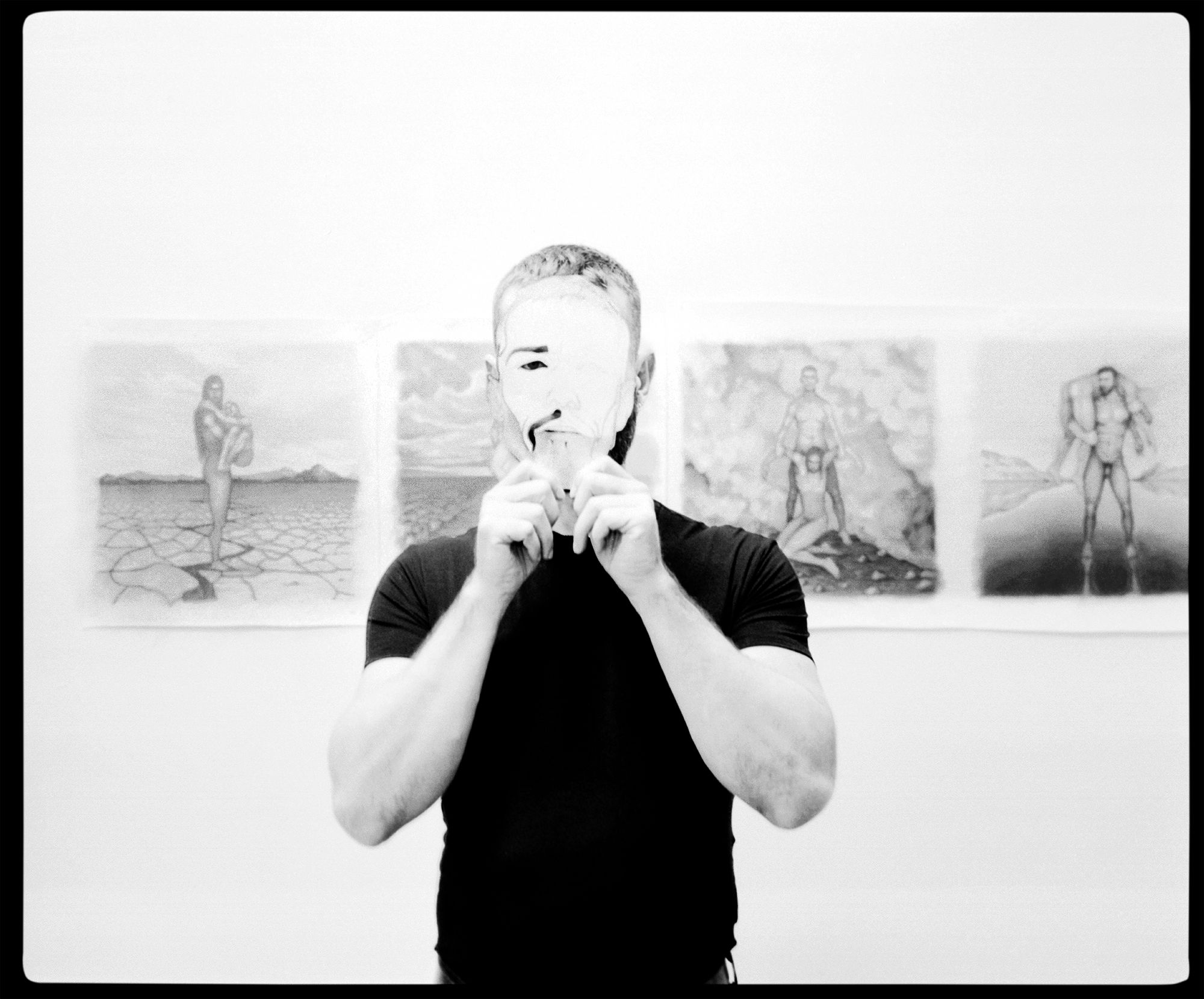 Carlos Motta standing in front of a wall of drawings, holding a mask up to his face