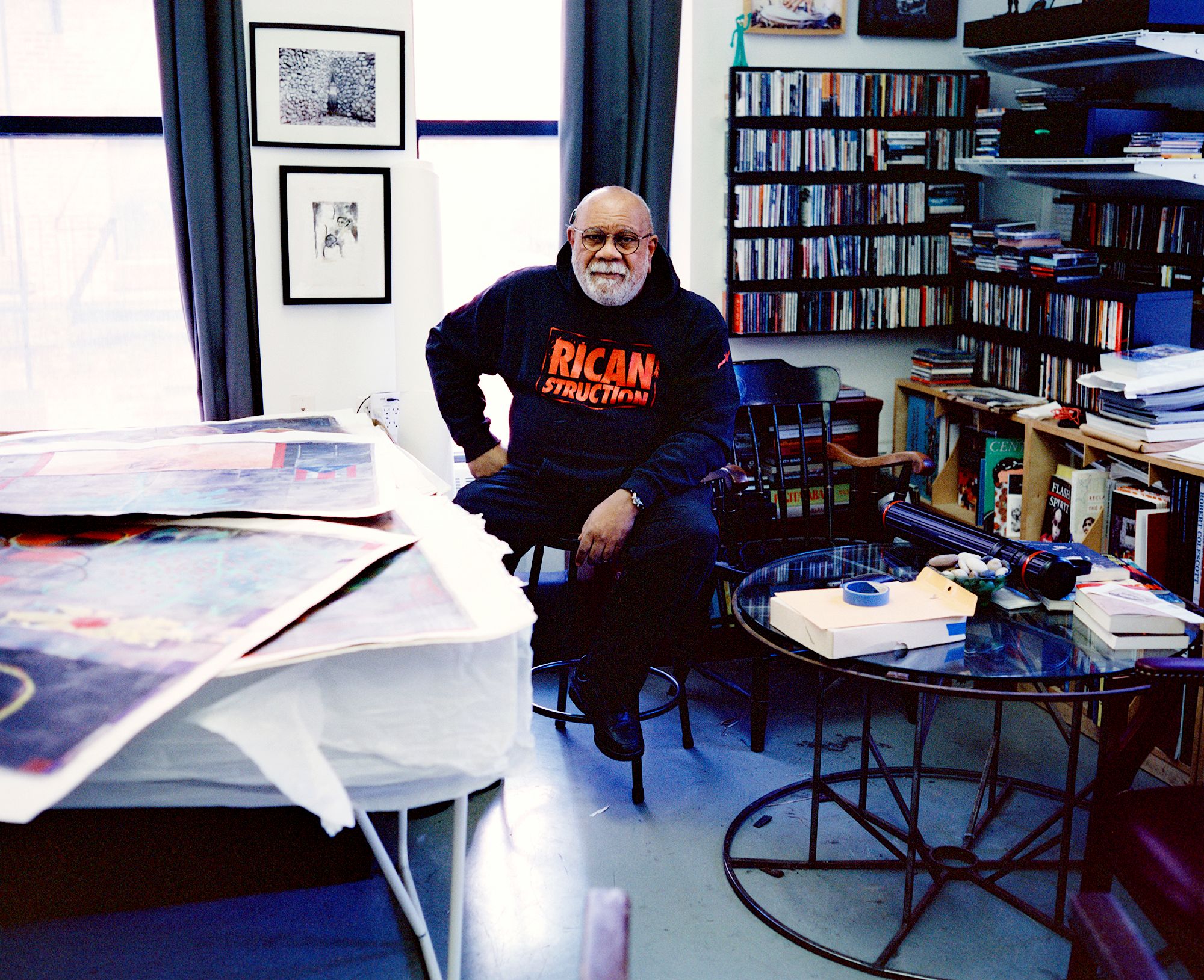 Juan Sánchez seated in his studio between tables covered with prints and books