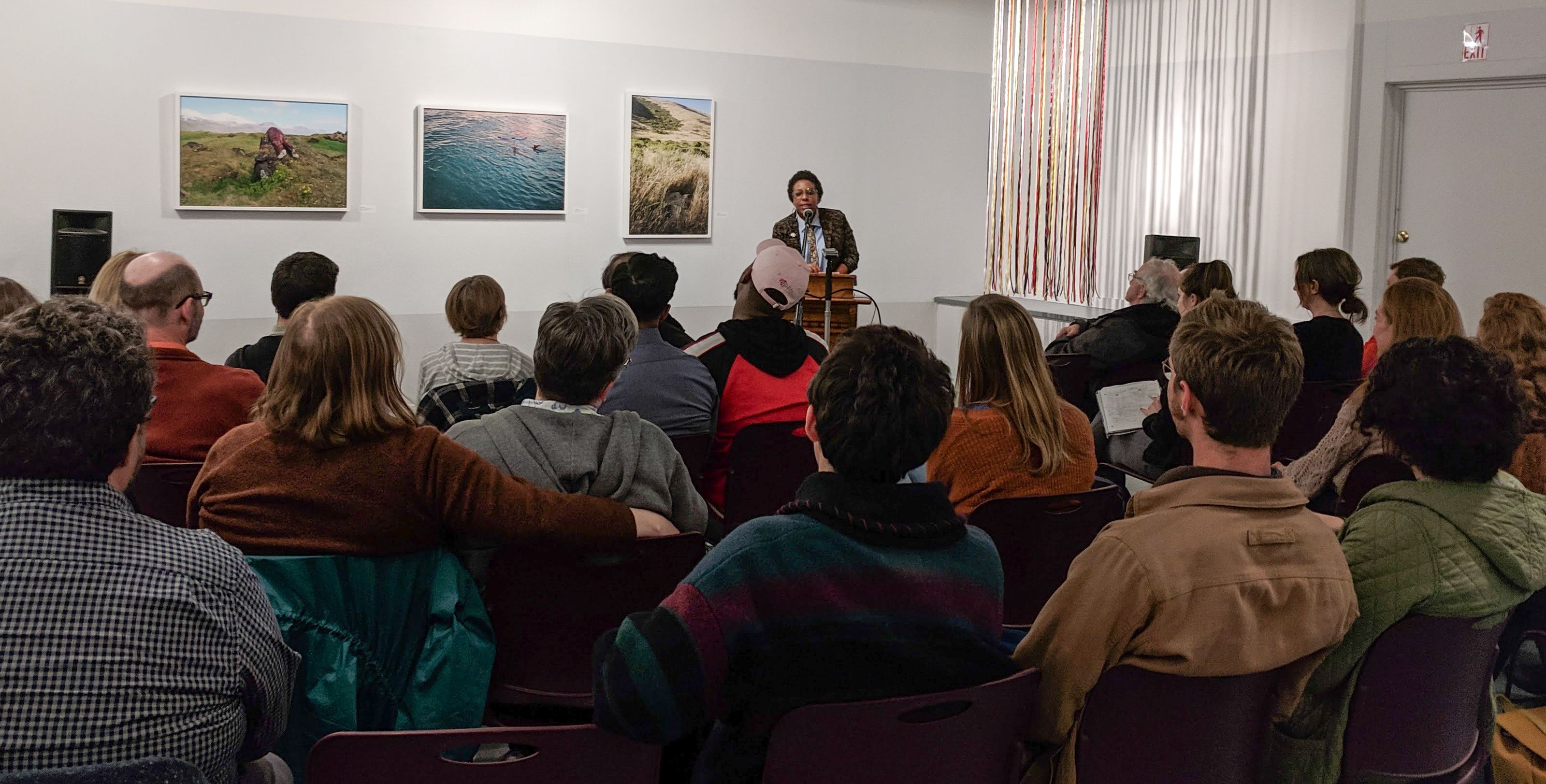 Man reading at a podium aloud to an audience. Artwork on the walls in the background.